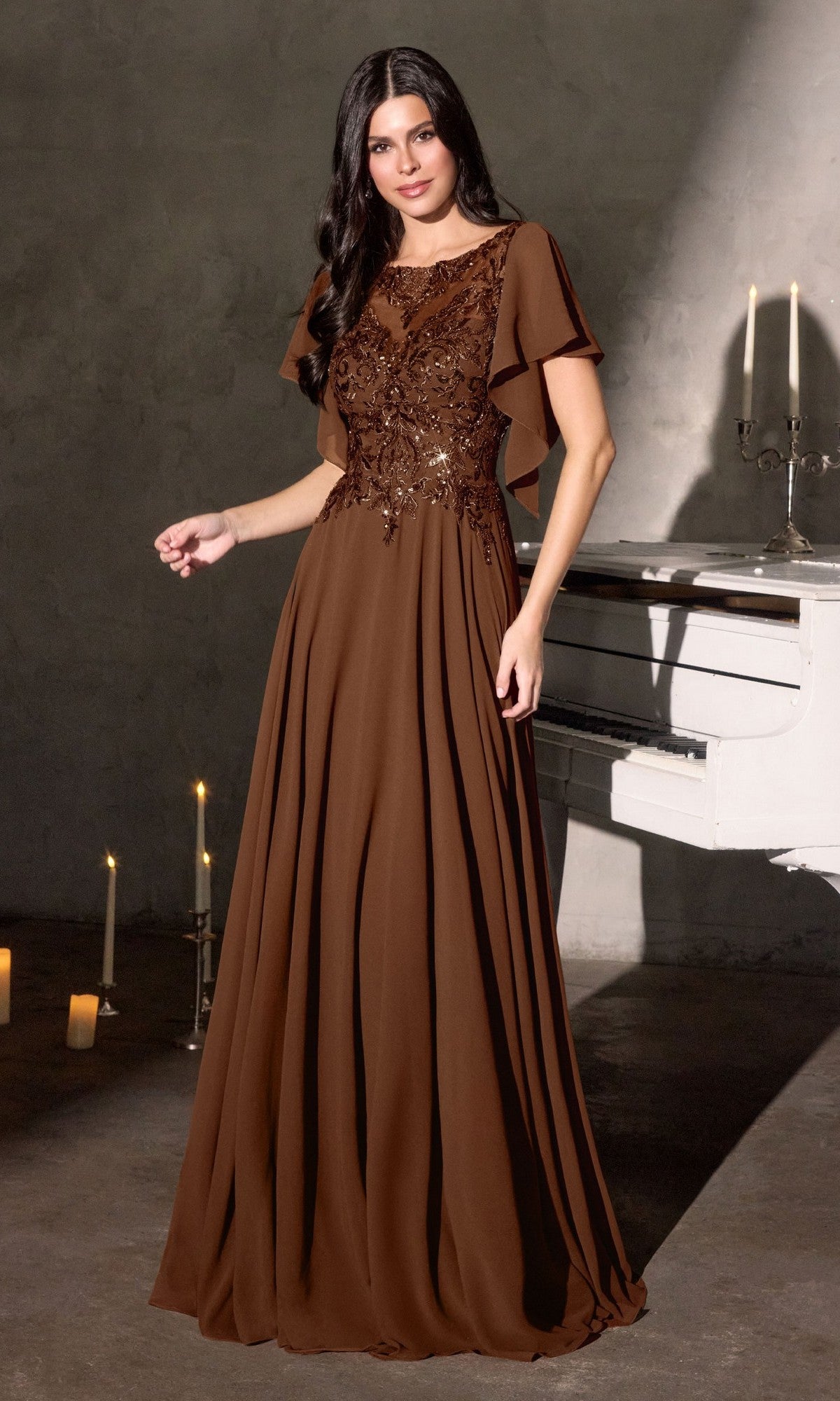 A woman in the Ladivine CL09 Long Formal Dress, a floor-length brown gown with an embroidered bodice and flutter sleeves, stands indoors near a white piano and lit candles.